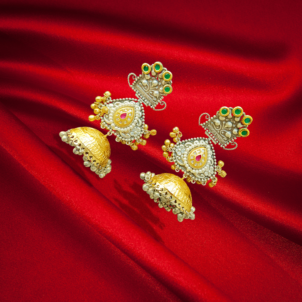 A pair of dual tone brass earrings with gold and silver plating, featuring intricate designs and high quality stones, displayed on a red fabric background.